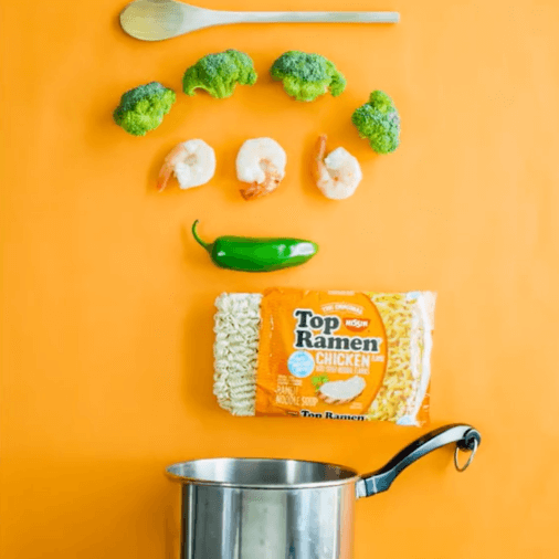 Wooden spoon, broccoli, shrimp and a jalapeno, Top Ramen package and pot lined up vertically on orange background.